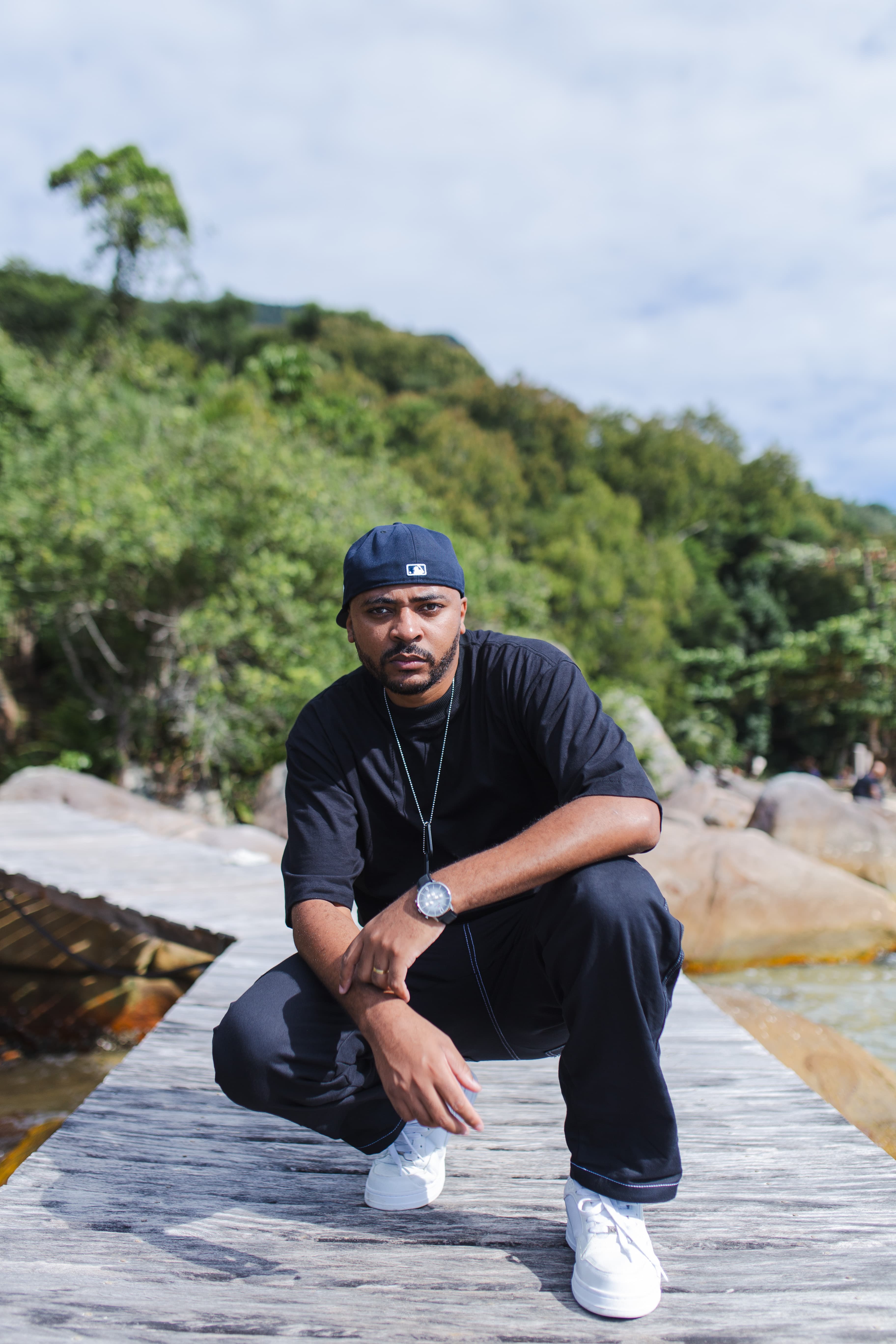 Denis MC agachado em cima de uma pedra fazendo pose de MC e olhando para a foto